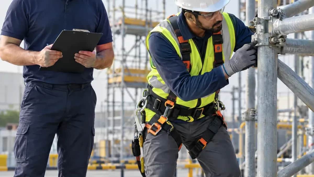 Safety supervisor with a clipboard watching a worker in a full-body harness check scaffold fittings on a construction site